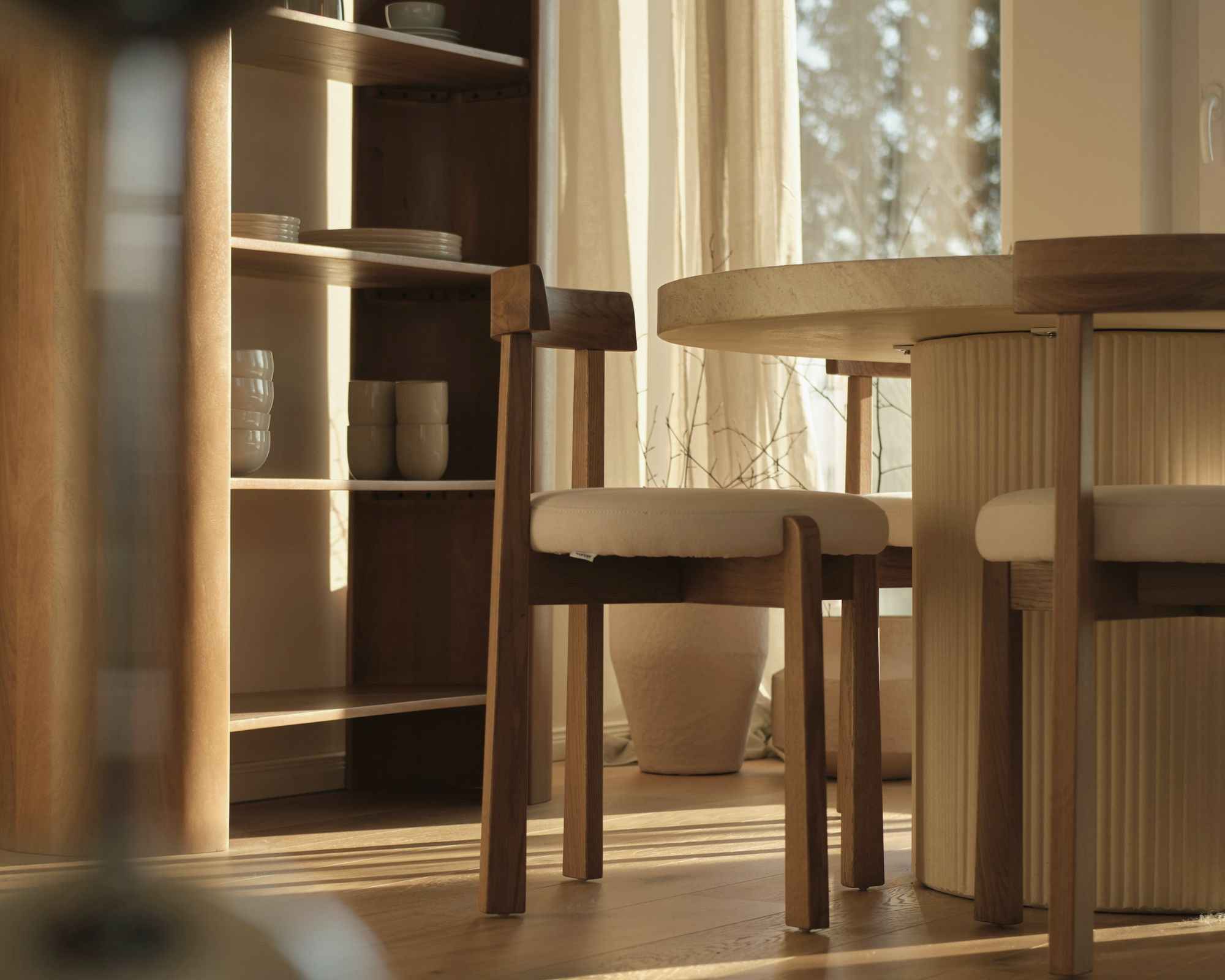 Late afternoon sun across an oak floor: a dining chair with a cream seat drawn up to a round table, open shelving stacked with plates and bowls, a linen curtain at the window.